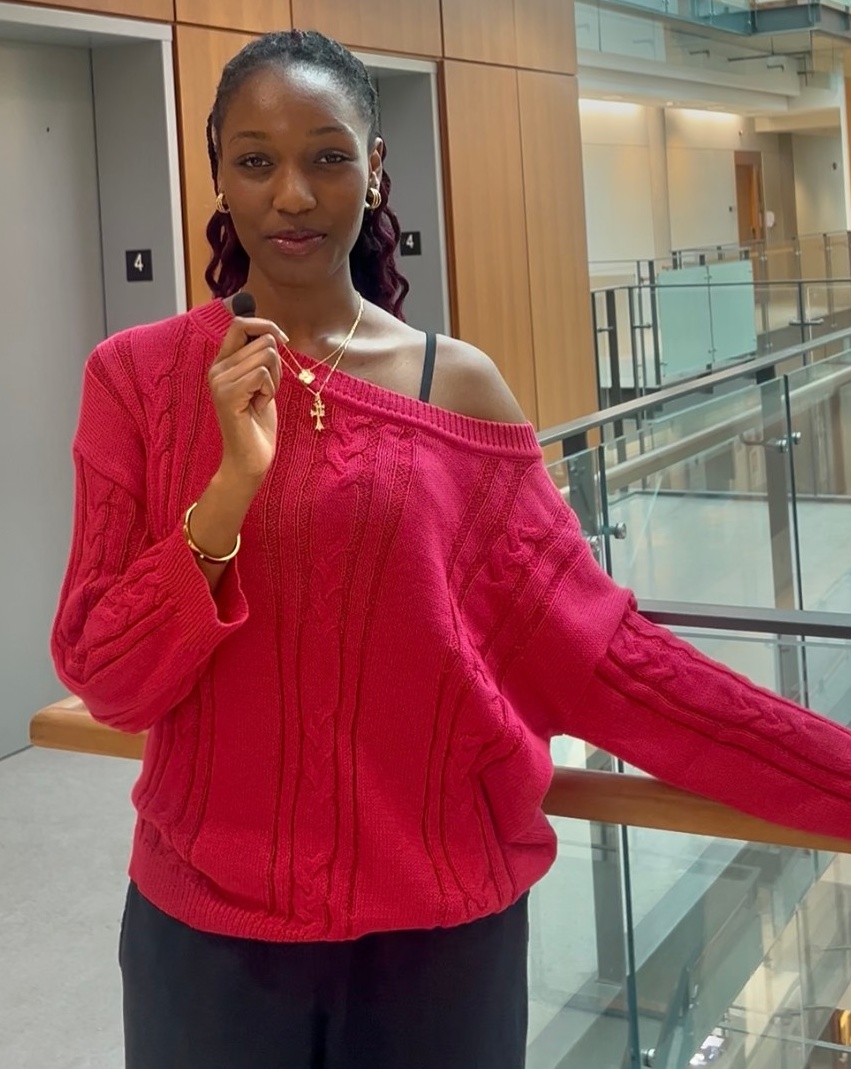 Jasmine stands in front of a balconey, holding a microphone and wearing a pink sweater.