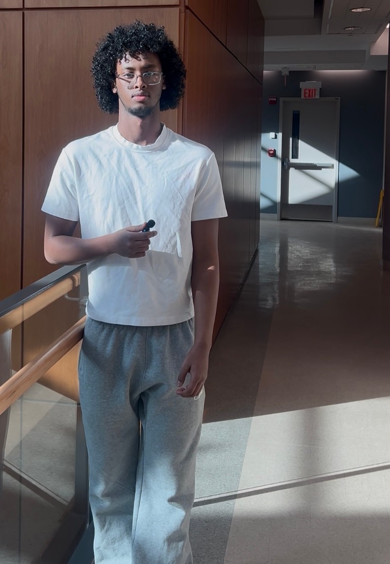 Mustafa stands next to a balconey, holding a microphone and wearing a white t-shirt.