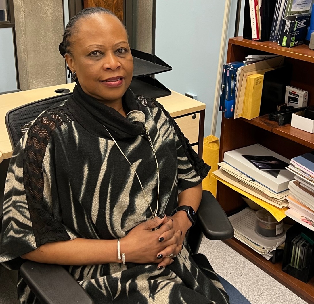 Joyline sits at her desk with her hands clasped, wearing a black and white sweater.