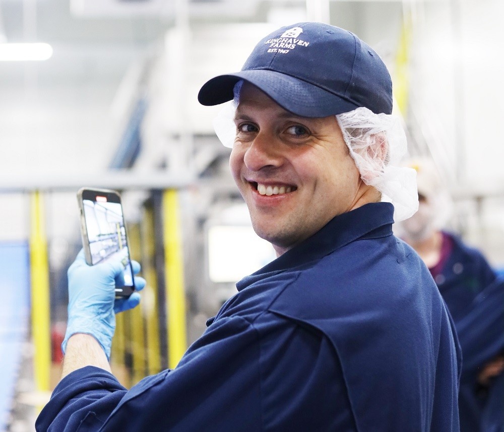 Jay, wearing a blue cap and work clothes, turns to smile at the camera. He is taking a photo of farm machinery.