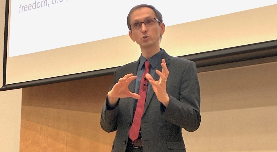 Dr. Gruzd, in grey suit and pink tie, gestures as he speaks on a stage.