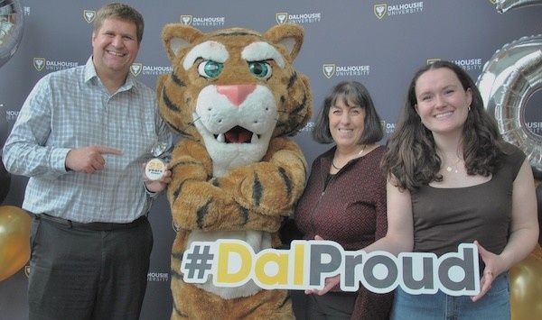 Brooklyn Malone stands against a Dal photo backdrop, with professors and the Dal Tiger mascot. She holds a sign that says "#DalProud"