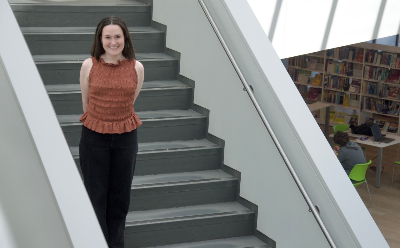 Brooklyn poses in a red shirt and black pants on the indoor stairs of the Halifax Central Library.