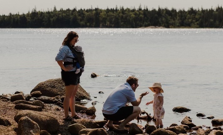 Nick and his family on a rocky beach. He crouches to talk to his small child, while his wife, carrying a baby, stands next to them. A dog swims in the water behind them.