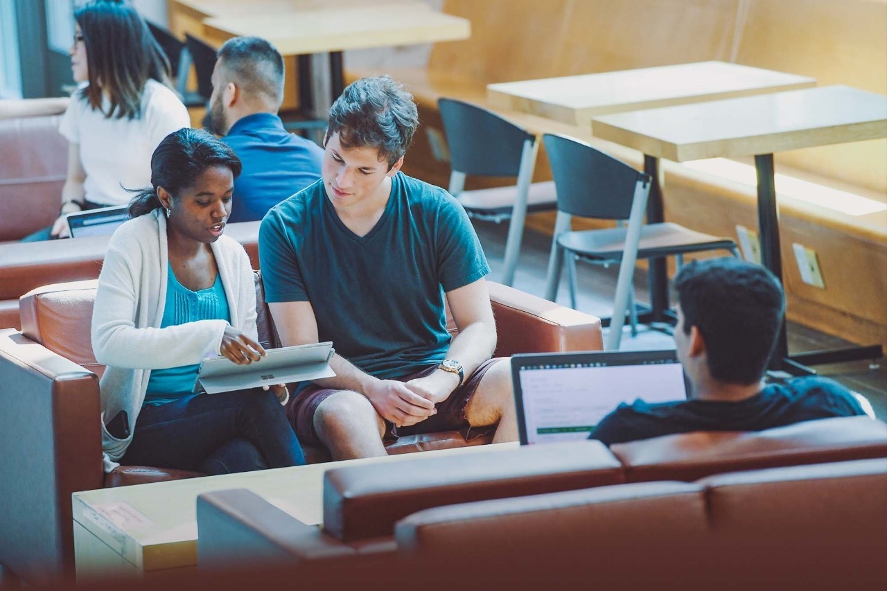 Students sit in a lounge area, consulting a tablet.
