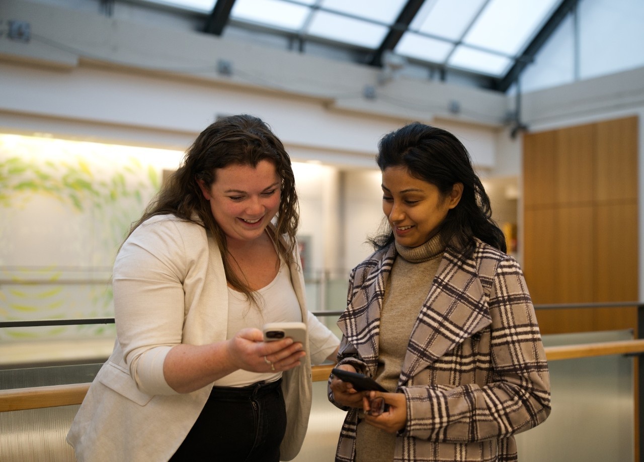 Two students smile at each other as they compare information on their phones. Two students smile at each other as they compare information on their phones.