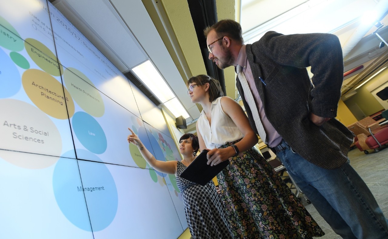 Students stand at a projection of data on a large wall, gesturing.