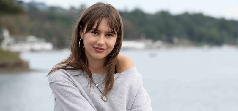 Abby sits on a rock facing the camera, smiling. In the background is water and the coastline.