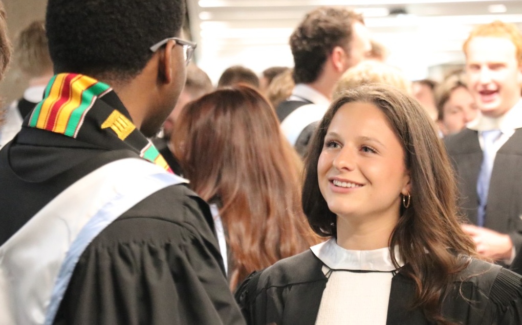 A student smiles at another student (facing away from camera). They are dressed in convcoation wear, with one wearing a kente sash in red, yellow, green and black.