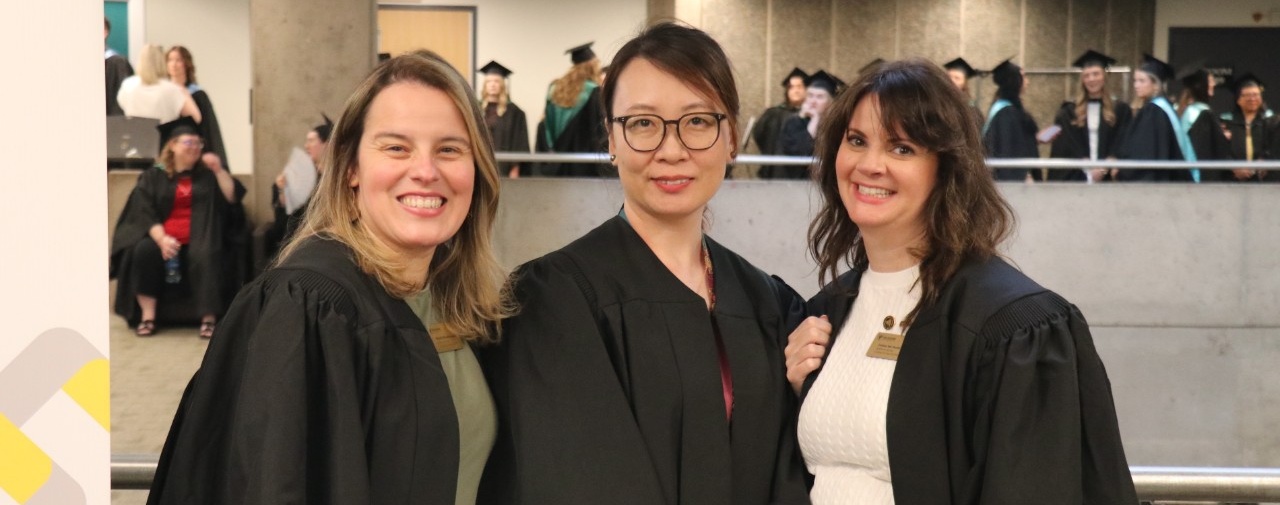 Three academic advisors dressed in black gowns smile at the camera. In the background, a group of students in robes preparing for their convocation ceremony.