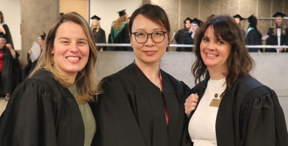 Three academic advisors dressed in black gowns smile at the camera. In the background, a group of students in robes preparing for their convocation ceremony.