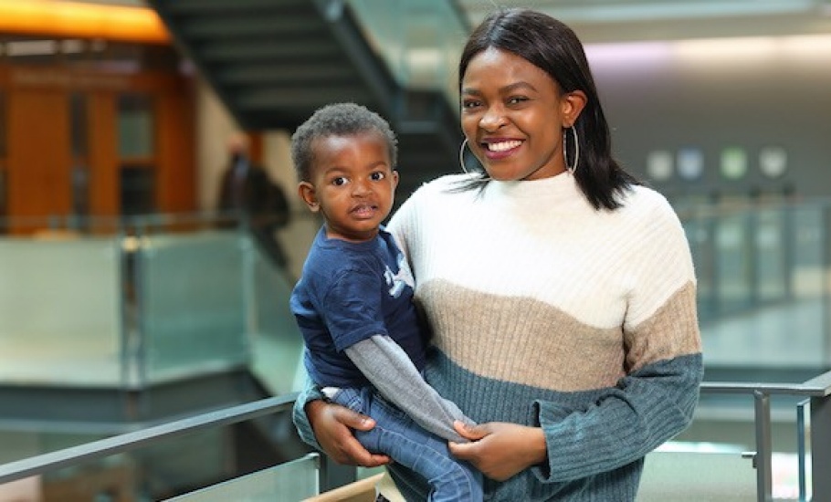 Rebecca Osamudiame, a Black woman with medium-length hair, stands next to a balcony in the Rowe building. She is wearing a white, blue and beige dress and gold hoop earrings. In one arm, she holds her son, a Black toddler wearing jeans and a blue shirt. She holds his hand with her free arm, and they both look at the camera, smiling.