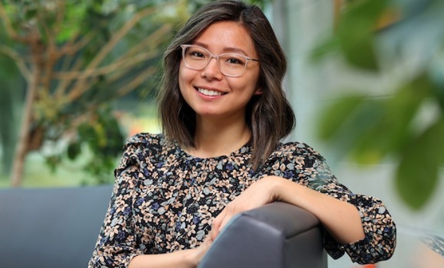 Byungmin Kang, a Korean woman with medium-length brown hair featuring light highlights, sits sideways on an armchair in the lobby of the Rowe building in front of a panel window with trees visible in the background. She is wearing a floral top and glasses, and smiling at the camera with one arm rested on the back of the armchair in front of her.