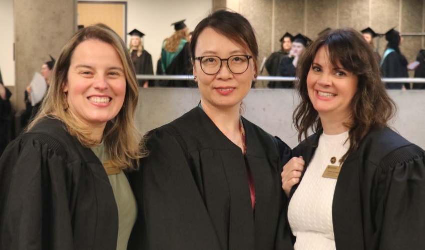 Three academic advisors dressed in black gowns smile at the camera. In the background, a group of students in robes preparing for their convocation ceremony.