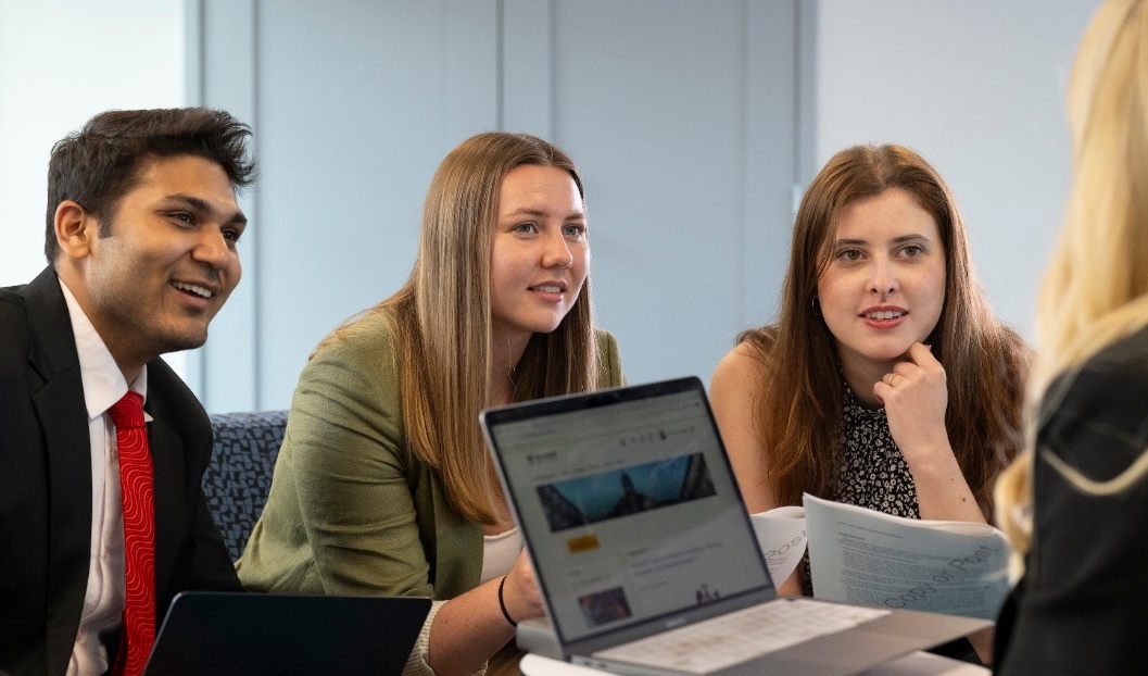 Three students dressed professionally sit on a bench, looking intently at someone using a laptop with their back to the camera.