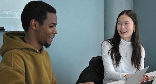 Two students sit at a table together in conversation, with laptops open in front of them, smiling.