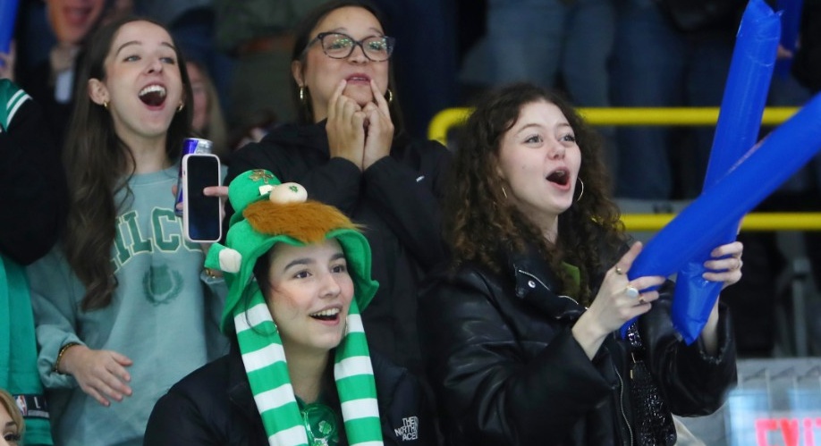 Four students standing in the seating area of an arena, smiling and cheering at something off-screen.