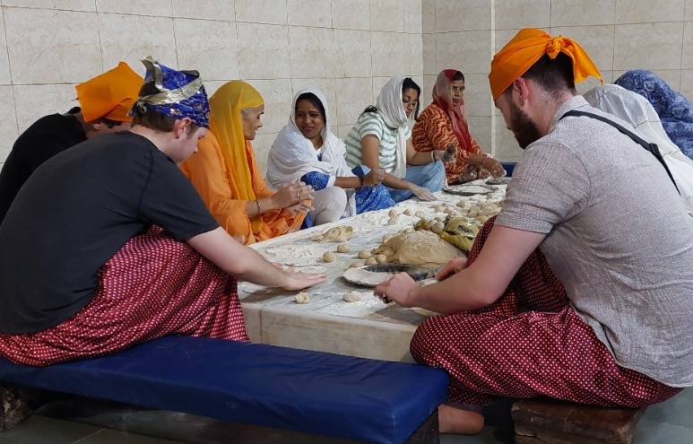 People in loose, brightly-coloured clothing sit cross-legged around a low table, talking and eating.