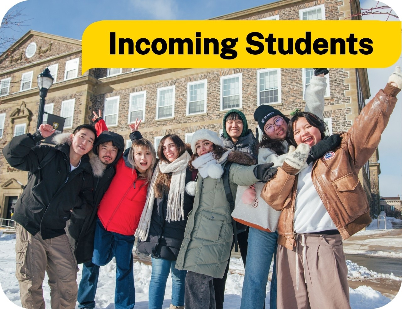 A group of students in winter clothing posing and smiling in front of a brick building.