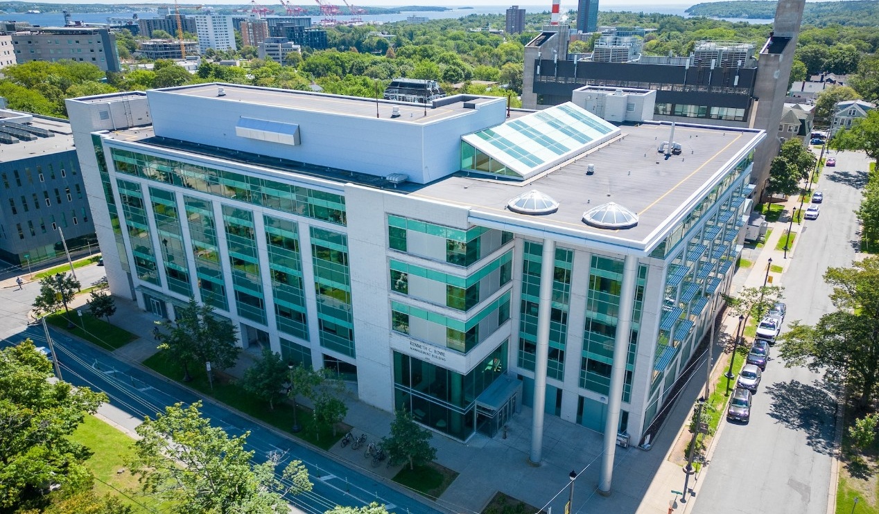 An aeiral shot of a green and grey building with many windows, surrounded by trees and other buildings.