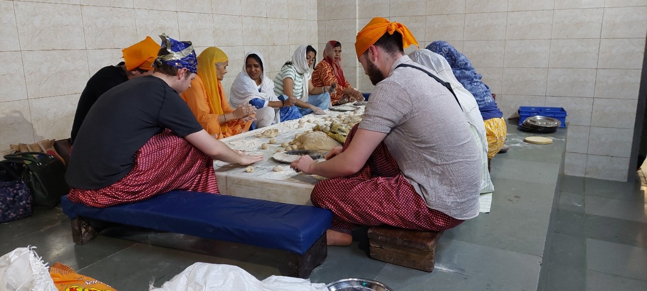 People in loose, brightly-coloured clothing sit cross-legged around a low table, talking and eating.
