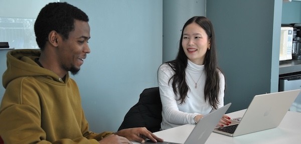 Two students sit at a table together in conversation, with laptops open in front of them, smiling.