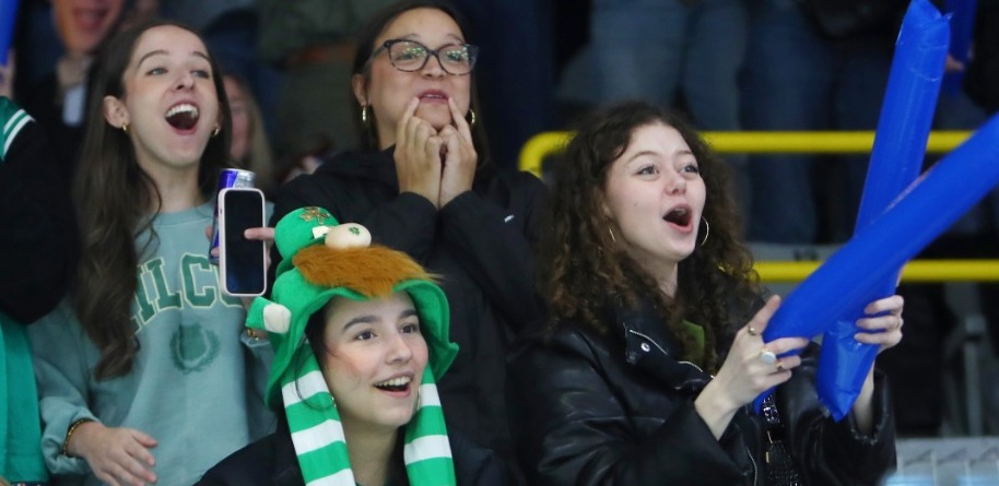Four students standing in the seating area of an arena, smiling and cheering at something off-screen.