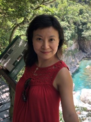 A photograph of Nan Geng, MA 2004, on a wood walking bridge in a wooded area by a sunny river.