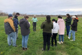 A group of students wearing jackets stand in a circle on a grassy field in springtime, listening to a woman at the centre of the circle speaking.