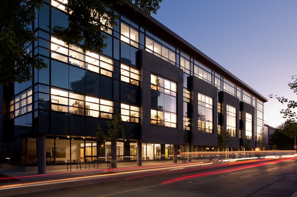 Pictured at dusk, the glass windows and concrete pillars of the Mona Campbell building are lit up by its interior lights. Traffic moves quickly past on the road outside.
