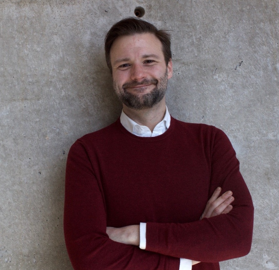 Andrew smiles. He has a light skin tone, short brown hair and dark stubble, and is wearing a dark red long-sleeved shirt.