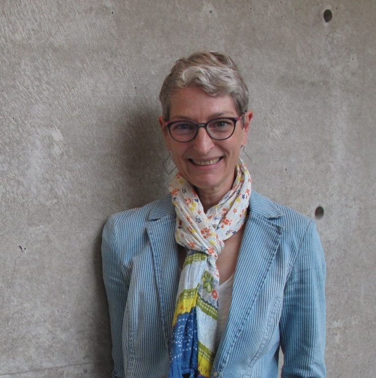 Georgia smiles at the camera. She has light-toned skin, short-cropped grey hair, and glasses, and she is wearing a blue jacket and colourful scarf.