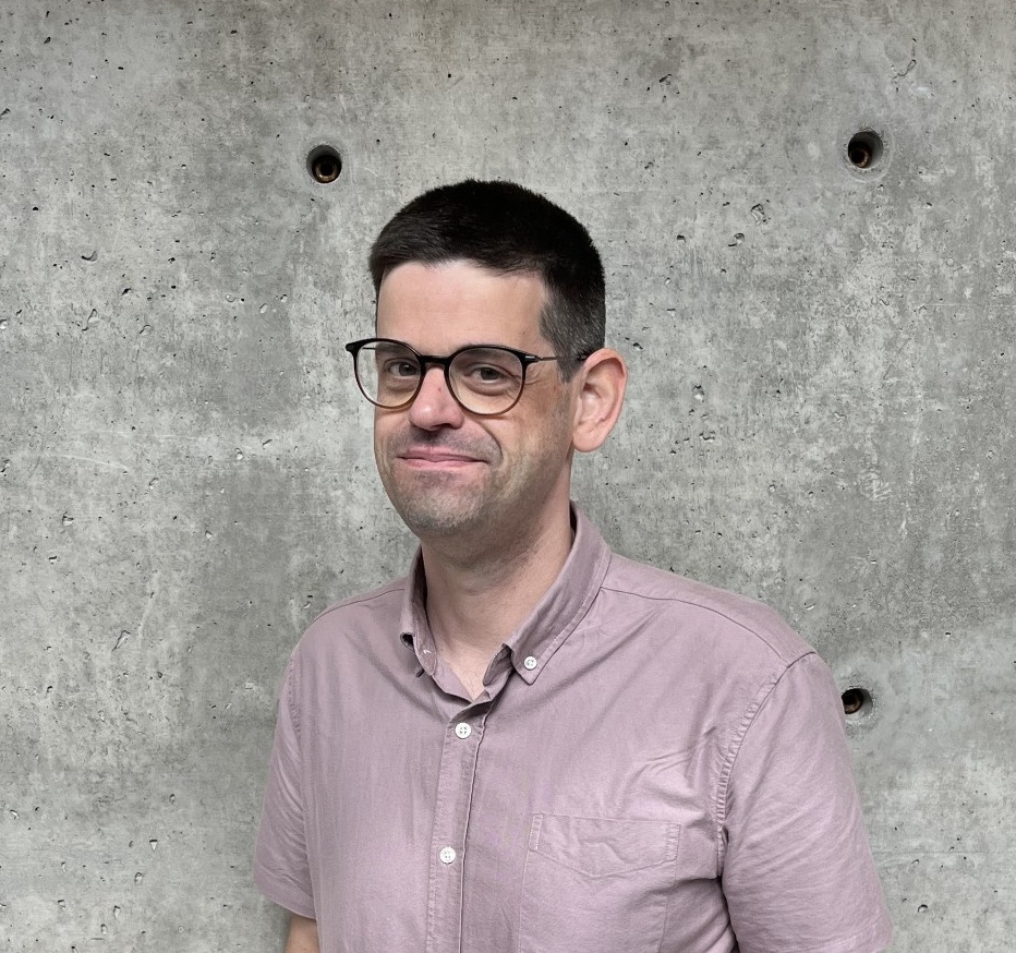 Will Langford smiles. He has a light skin tone and short brown hair, and is wearing glasses and a pink buttoned shirt.