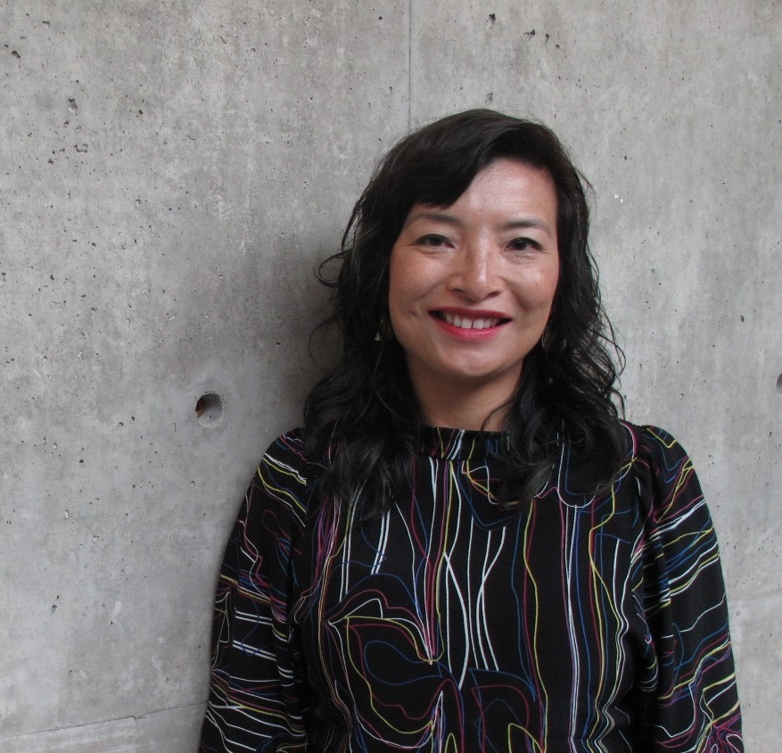 San smiles. She has a light skin tone and shoulder-length black hair, and is wearing a black shirt with lots of multi-coloured lines woven across.