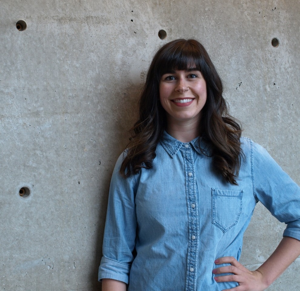 Laurel smiles. She has a light skin tone; long, dark-brown hair; and a nose piercing with a small gold ring. She is wearing a light blue shirt.