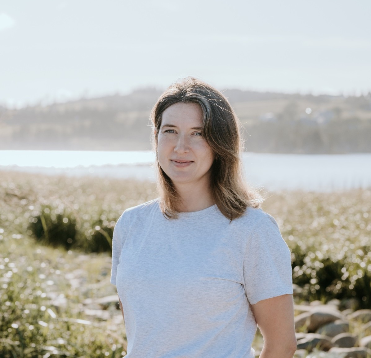 Phoebe is standing outdoors on a sunny day, smiling. She has a light skin tone and shoulder-length brown hair, and is wearing a white t-shirt.