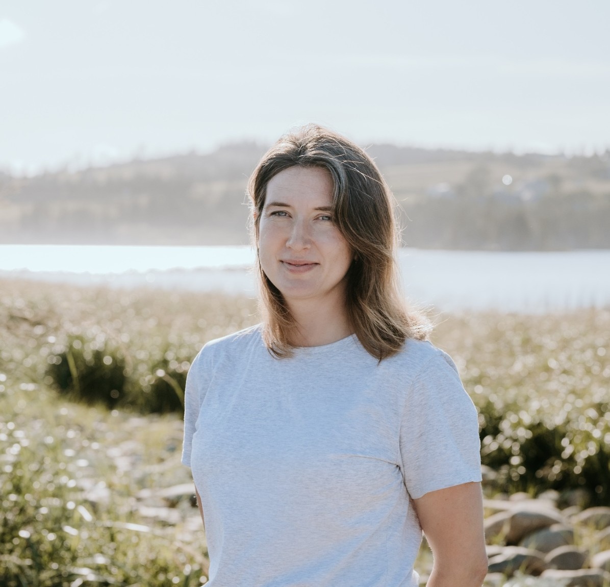 Phoebe is standing outdoors on a sunny day, smiling. She has a light skin tone and shoulder-length brown hair, and is wearing a white t-shirt. Phoebe is standing outdoors on a sunny day, smiling. She has a light skin tone and shoulder-length brown hair, and is wearing a white t-shirt.