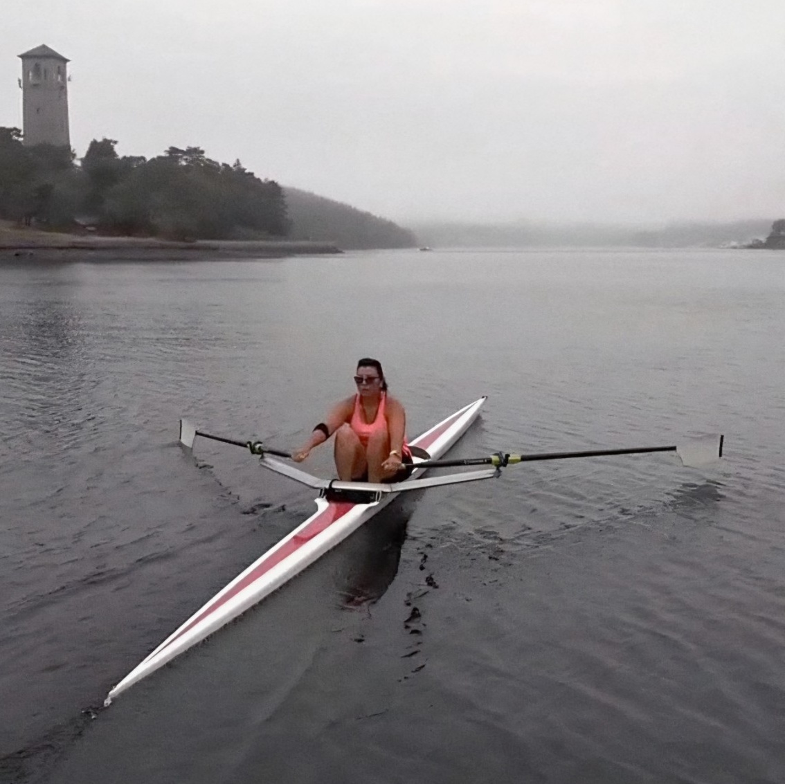 On a foggy day, San rows a small, thin boat across the empty north-west arm of Halifax Harbour. Trees and the Dingle Tower are visible on the shoreline nearby.