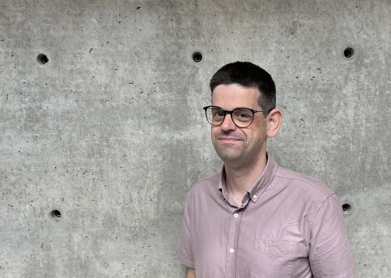 Will smiles at the camera. He has a light skin tone, short dark hair, and glasses.