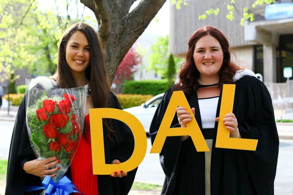 Two smiling graduates stand outside on a sunny day, dressed in black convocation gowns. One has a bouquet of red roses resting on her arm, and together the grads are holding up three large yellow letters that spell “DAL”.