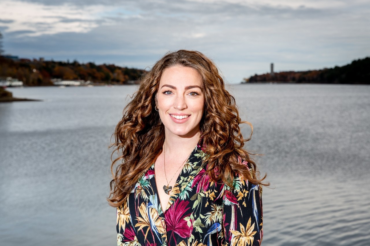 Alexa Goodman smiles at the camera. They have long, reddish-brown hair and a shirt with multi-coloured flowers on it. They are standing in front of a harbour.