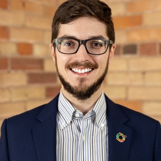 Lars smiles. He has a light skin tone, short brown hair and a tidy beard and moustache. He is wearing glasses and a dark blue jacket over a striped shirt.