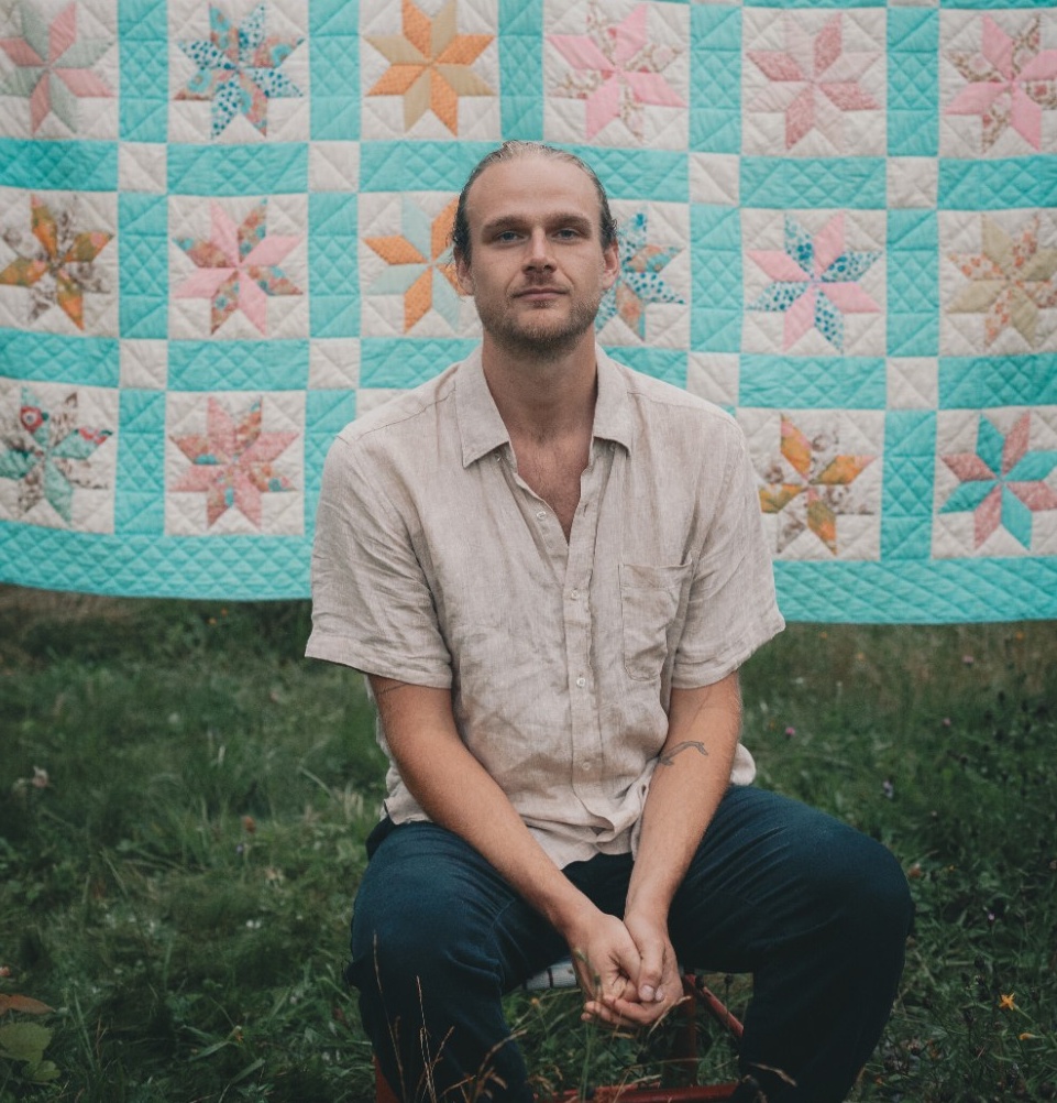 Alex smiles. He has a light skin tone, long brown hair tied back, and light stubble. He is wearing a cream-coloured shirt and blue jeans, and is sitting outdoors on a lawn in front of a hung-up quilt with colourful star patterns.