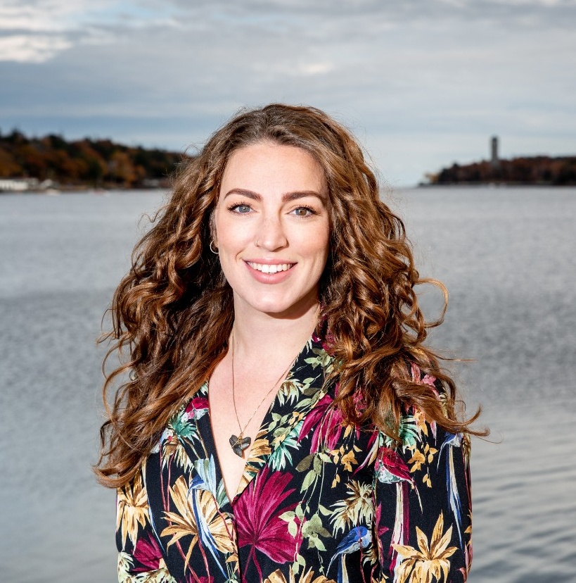 Alexa smiles. They have a light skin tone and long, reddish-brown hair, and are wearing a black shirt with multi-coloured flowers on it. They are standing in front of a harbour under a cloudy sky.