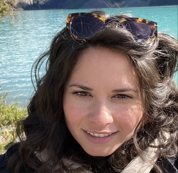 Simone smiles at the camera, standing outdoors on a sunny day. She has a light skin tone; long, dark, wavy hair; and a pair of sunglasses perched on her head.