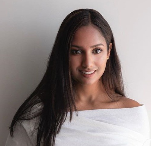 Divya smiles at the camera. She has a medium skin tone and long dark hair, and she is wearing a white shirt.