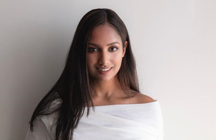 Divya smiles at the camera. She has a medium skin tone and long dark hair.