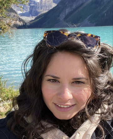 Simone smiles at the camera, standing outdoors on a sunny day. She has a light skin tone; long, dark, wavy hair; and a pair of sunglasses perched on her head.
