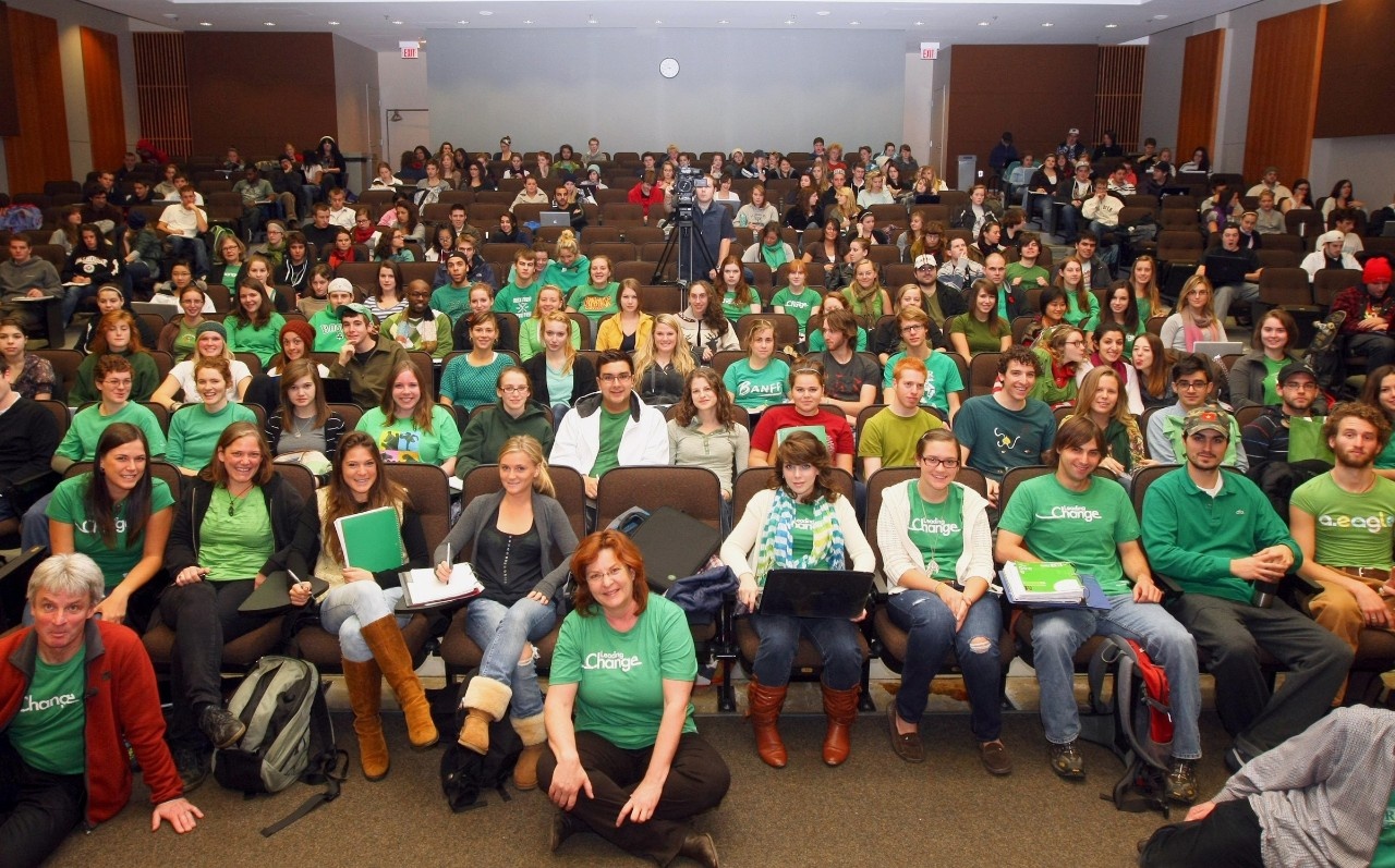 Group of 80-100 smiling students seated in the Ondaatje Auditorium wearing green "Leading Change" t-shirts.