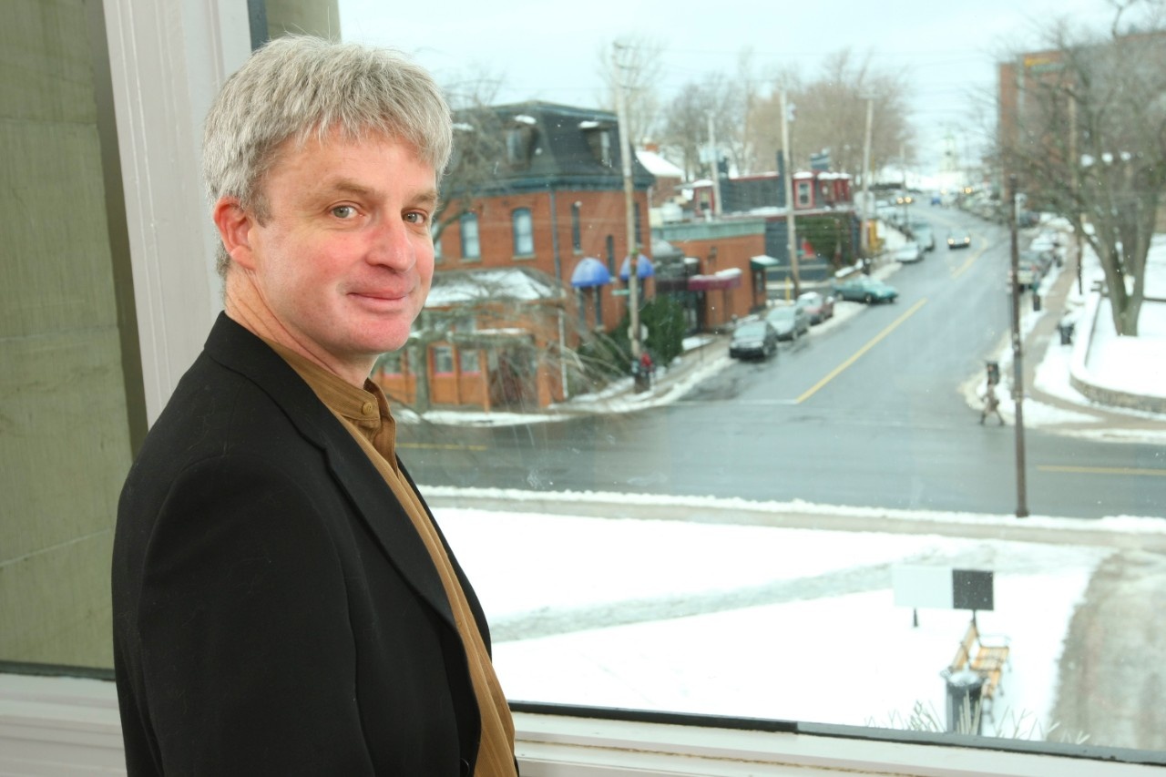 Steve stands next to a window that looks out onto a snowy Halifax street. He has a light skin tone and short greying hair, and is wearing a black jacket over a brown shirt.